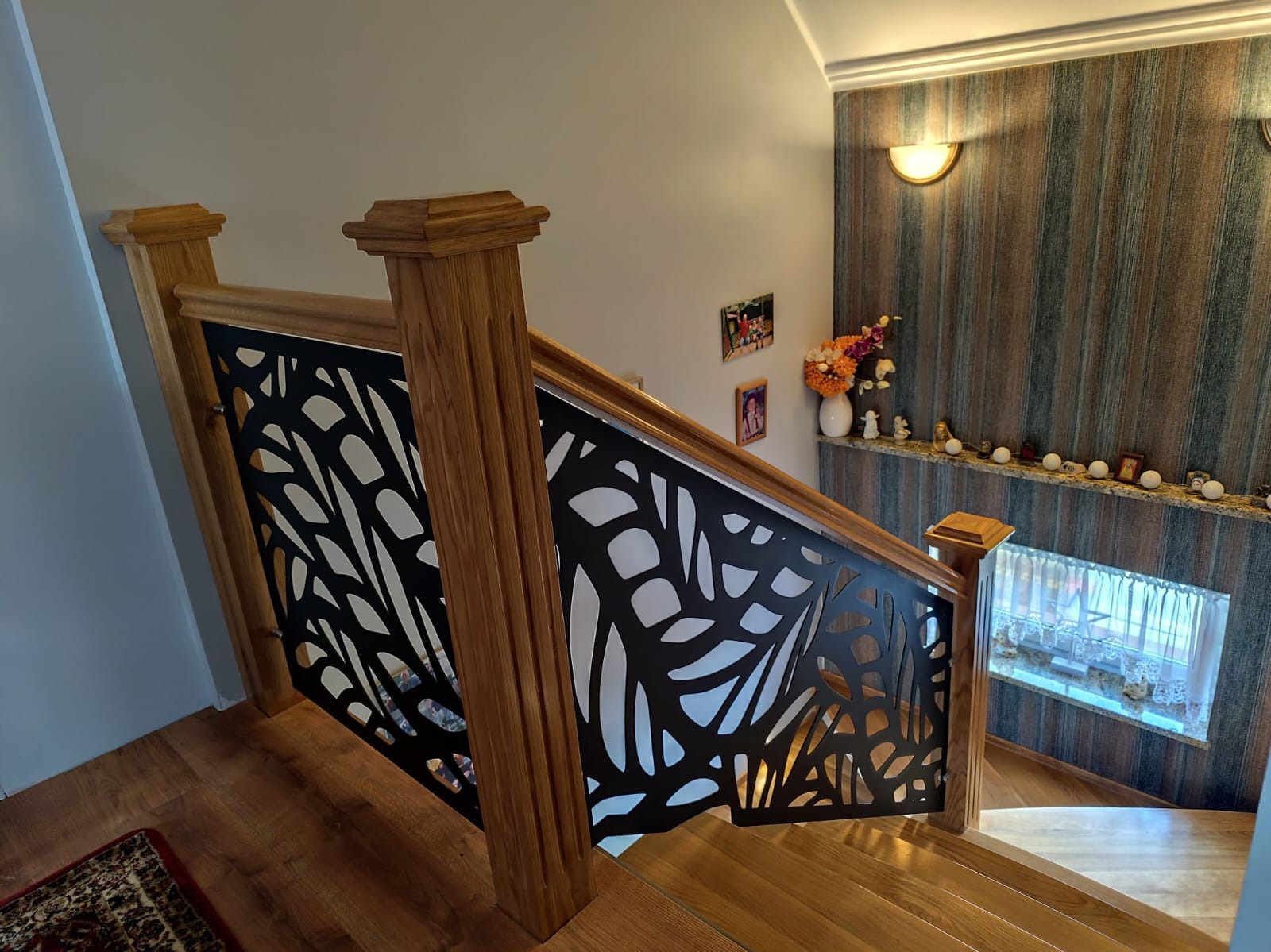 Drewniana balustrada schodowa z czarnym, ażurowym wypełnieniem o motywie liściastym. Widok z góry na schody z drewnianymi stopniami i dekoracyjną ścianą w tle.