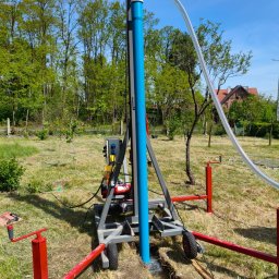 EKOSTUDNIA ARTUR BENTKOWSKI - Wiertnica do studni głębinowych w trakcie pracy na zewnątrz. Widoczny sprzęt, niebieska rura w ziemi, wykop i trawa. W tle drzewa i budynek.