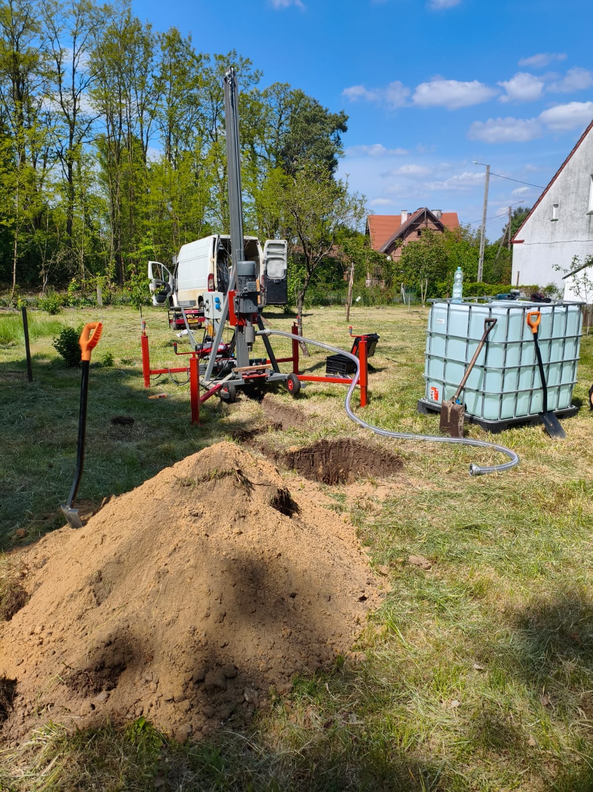 Wiertnica w ogrodzie podczas pracy. Widoczny zbiornik na wodę, wykopane otwory i sterta ziemi. W tle dom i samochód dostawczy.