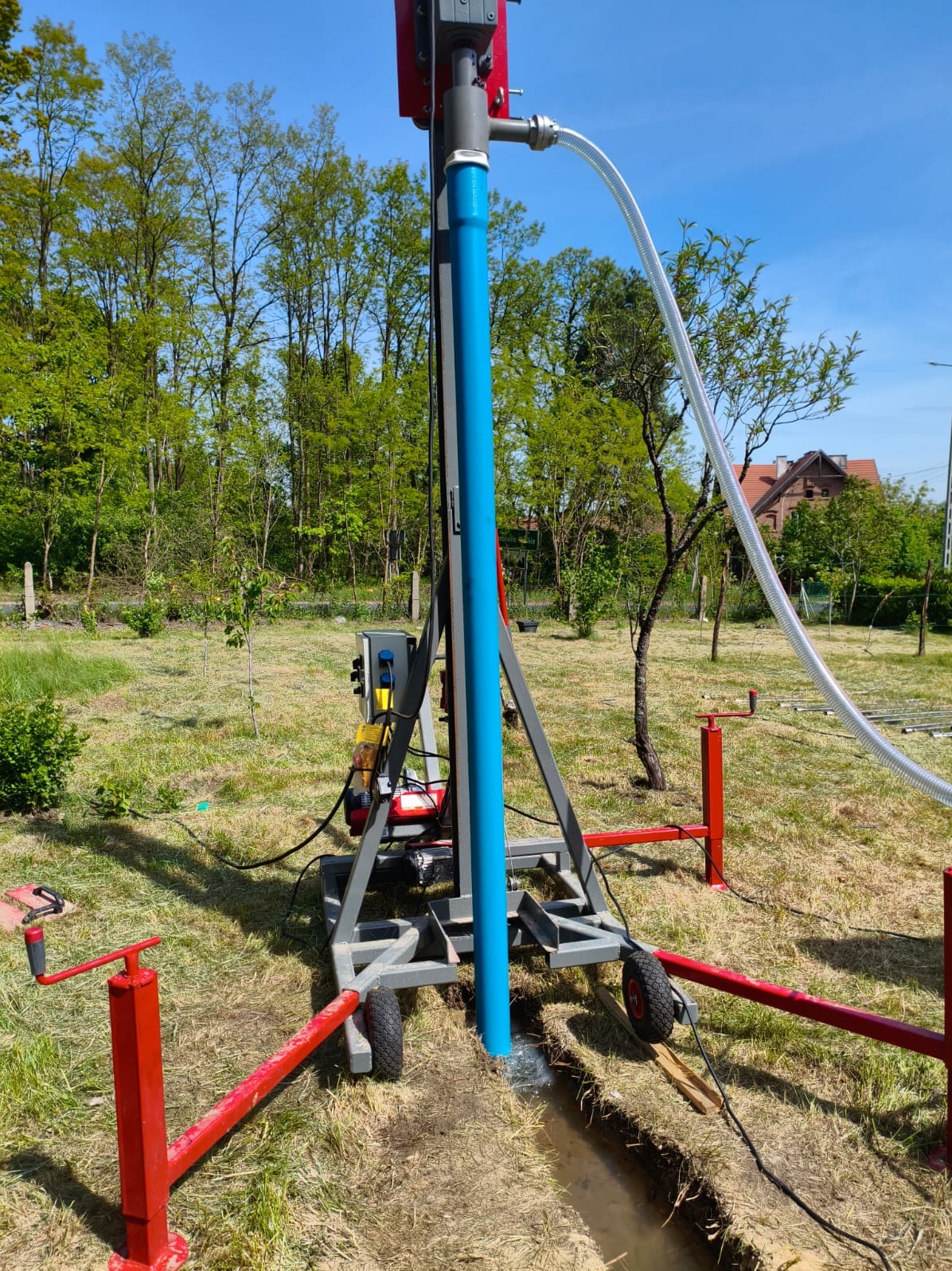 Wiertnica do studni głębinowych w trakcie pracy na zewnątrz. Widoczny sprzęt, niebieska rura w ziemi, wykop i trawa. W tle drzewa i budynek.