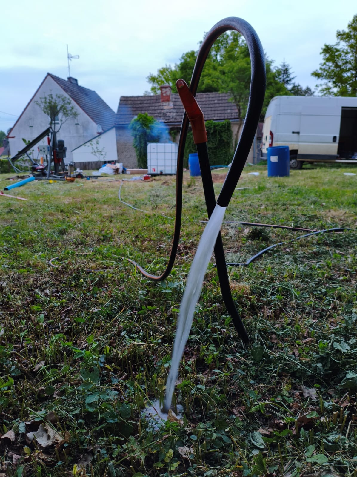 Woda wypływająca z węża na trawniku, w tle dom, sprzęt wiertniczy i biały van. Test wydajności po wierceniu.