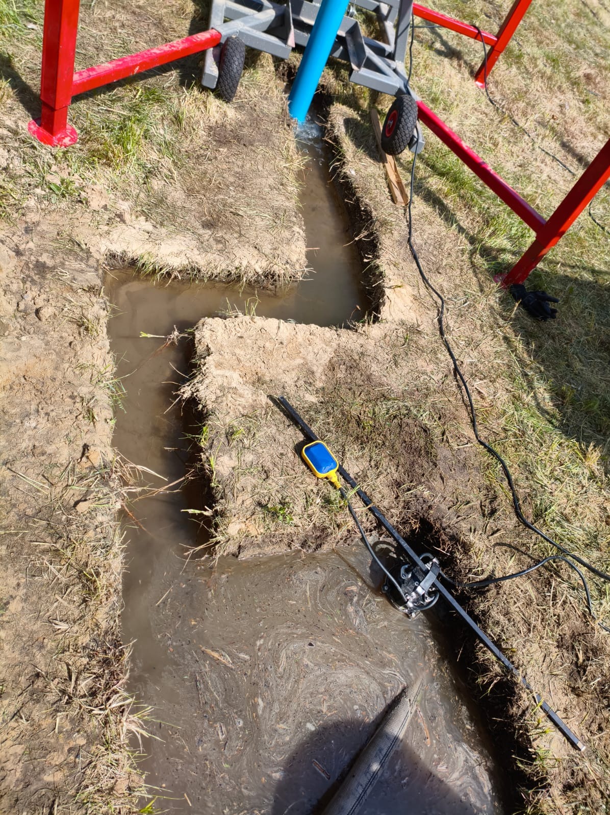 Wykop wypełniony wodą podczas wiercenia studni głębinowej. Widoczna pompa z pływakiem i fragment ramy wiertniczej. Prace ziemne w toku, teren trawiasty.