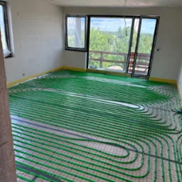 Zielone rury ogrzewania podłogowego ułożone na izolacji w przestronnym, jasnym pokoju z dużymi oknami wychodzącymi na zalesiony teren.