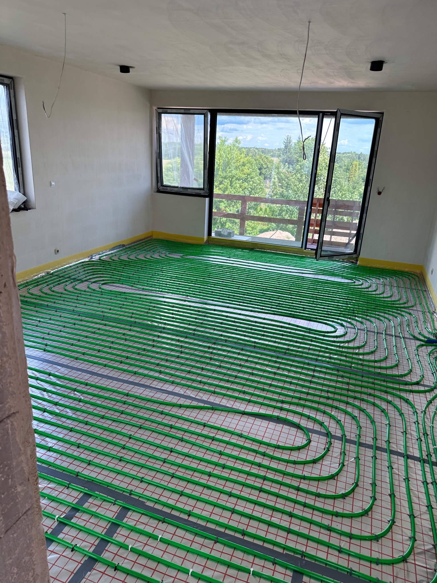 Zielone rury ogrzewania podłogowego ułożone na izolacji w przestronnym, jasnym pokoju z dużymi oknami wychodzącymi na zalesiony teren.