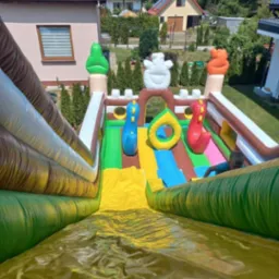 Widok z góry na dmuchaną zjeżdżalnię na trawniku z elementami dekoracyjnymi w kształcie lodów i dinozaurów, w tle domy jednorodzinne.