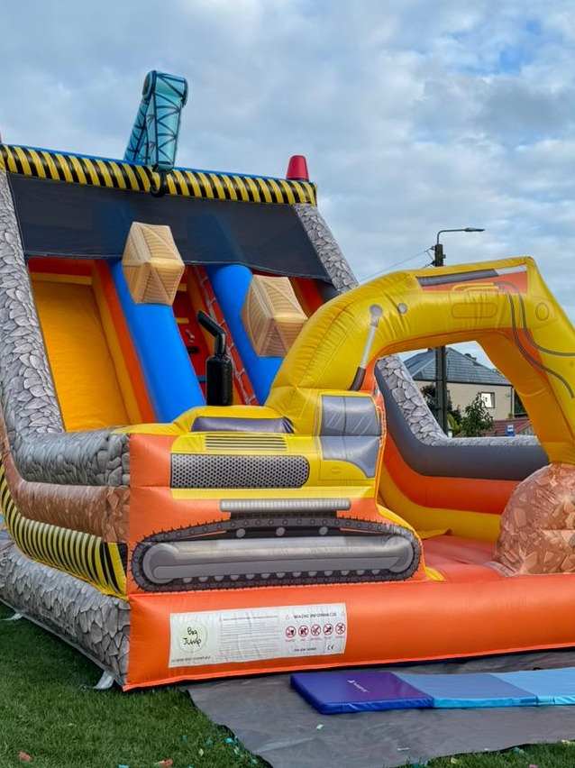 Duży, kolorowy dmuchany zamek w kształcie koparki z żółtymi i pomarańczowymi elementami, zjeżdżalnią i imitacją przeszkód, ustawiony na trawie.