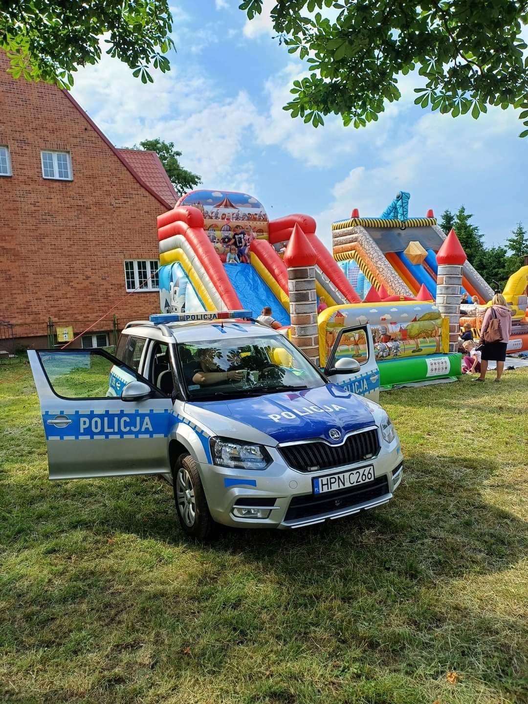 Radiowóz policyjny z otwartymi drzwiami na trawie, w tle dmuchane zamki i zjeżdżalnie w słoneczny dzień.