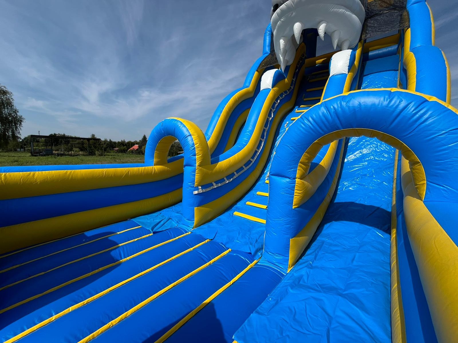 Niski kąt ujęcia na dużą, nadmuchiwaną zjeżdżalnię w kolorach niebieskim i żółtym, z motywem paszczy rekina na szczycie, umieszczoną na trawiastym terenie pod błękitnym niebem z lekkimi chmurami.