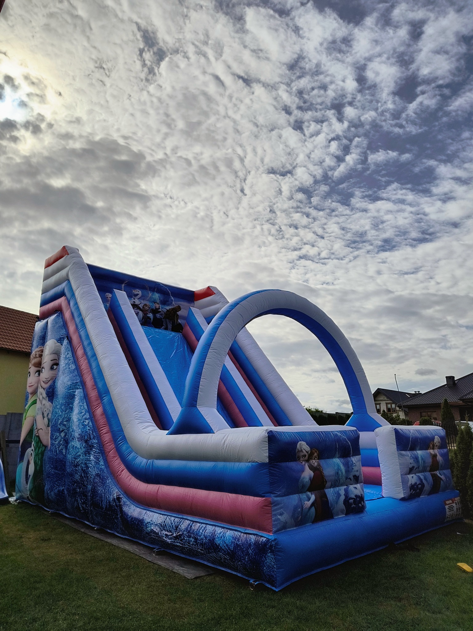 Duży, nadmuchiwany zamek-zjeżdżalnia z motywem 'Kraina Lodu' na trawniku przed domem, widoczne postacie Elsy i Anny, pochmurne niebo w tle.