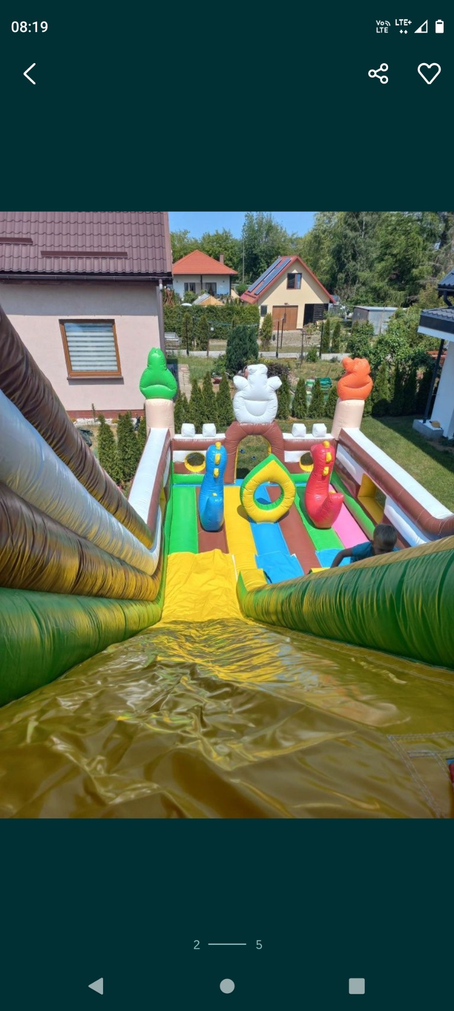 Widok z góry na dmuchaną zjeżdżalnię na trawniku z elementami dekoracyjnymi w kształcie lodów i dinozaurów, w tle domy jednorodzinne.