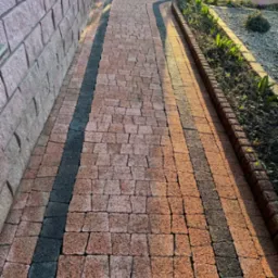 Kaskadowa ścieżka z kostki brukowej w odcieniach czerwieni i czerni, obrzeża rabat z roślinnością, mur z kamienia w tle, widok z góry, ciepłe światło.