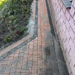 Dawid Zięba - Częściowo wyczyszczona kostka brukowa w odcieniach czerwieni i czerni, obok muru budynku i rabaty z roślinami. Na końcu ścieżki myjka ciśnieniowa.