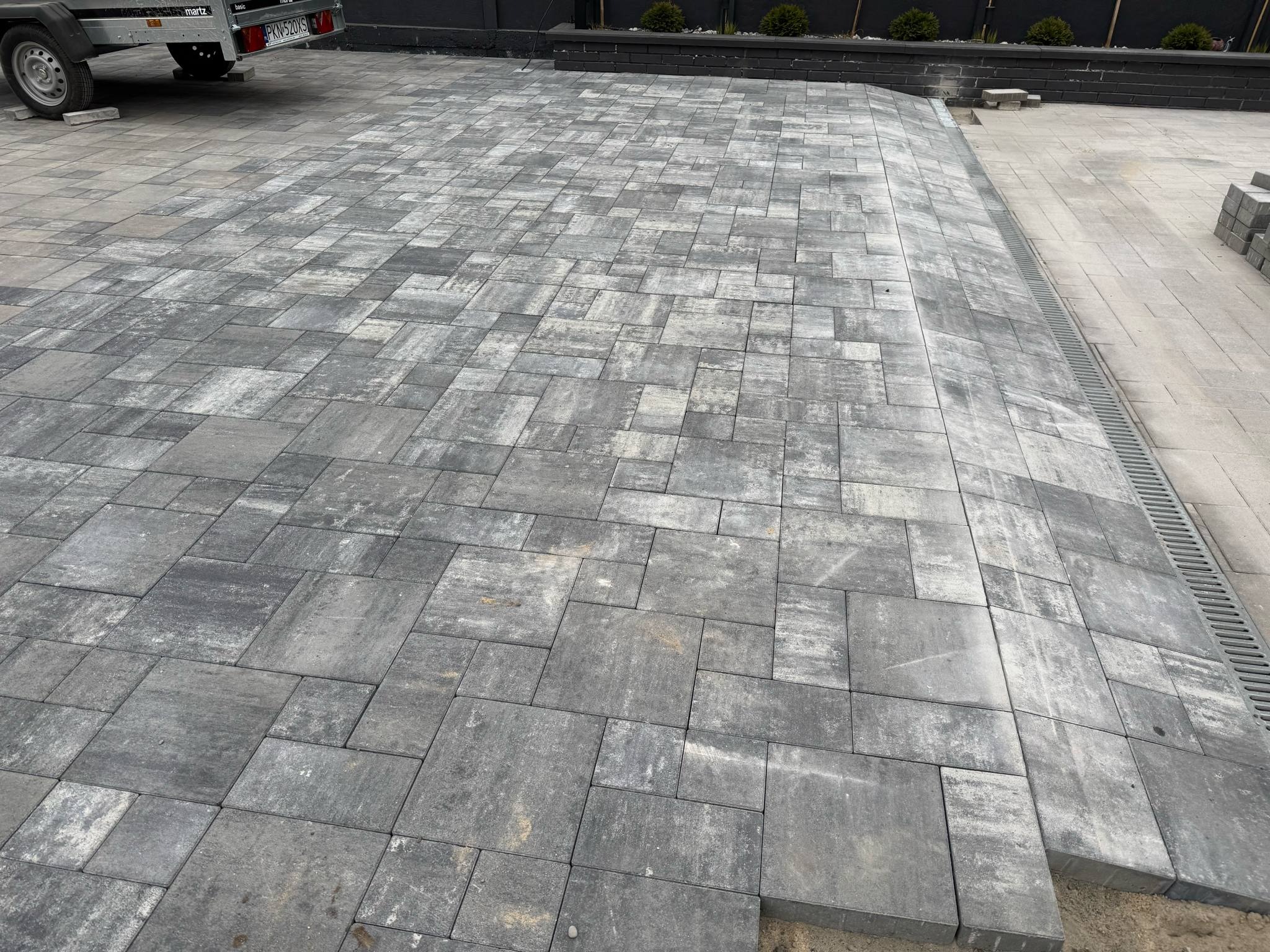 Ułożona, szara kostka brukowa na posesji z widoczną kratką odpływową. Częściowo widoczna przyczepka samochodowa i mur. Elementy kostki ułożone w nieregularny wzór.