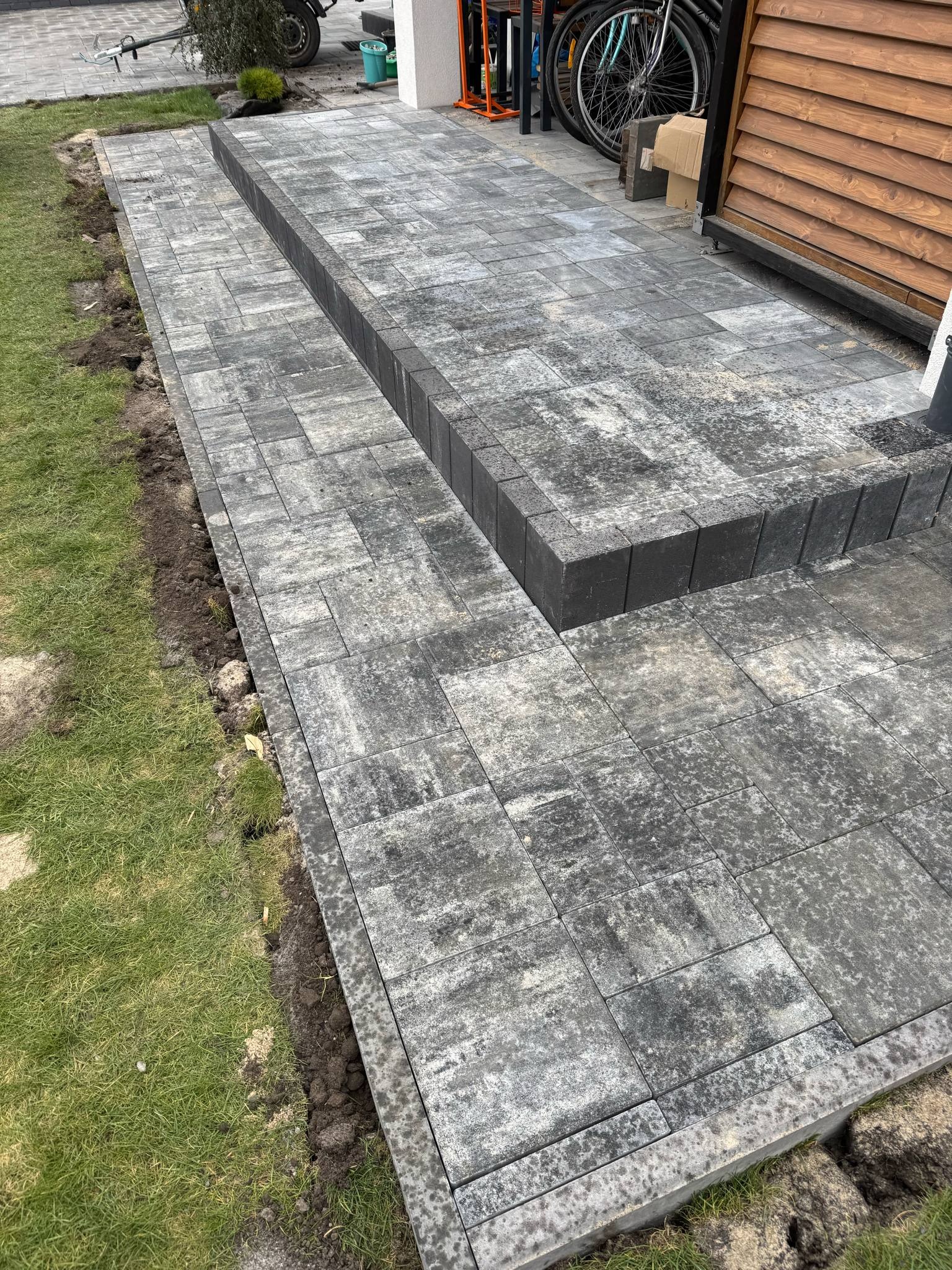 Ścieżka z szarej kostki brukowej z obrzeżem, prowadząca do drewnianego ogrodzenia. Widoczne rowery i elementy wyposażenia. Ujęcie z boku, z trawnikiem po lewej.