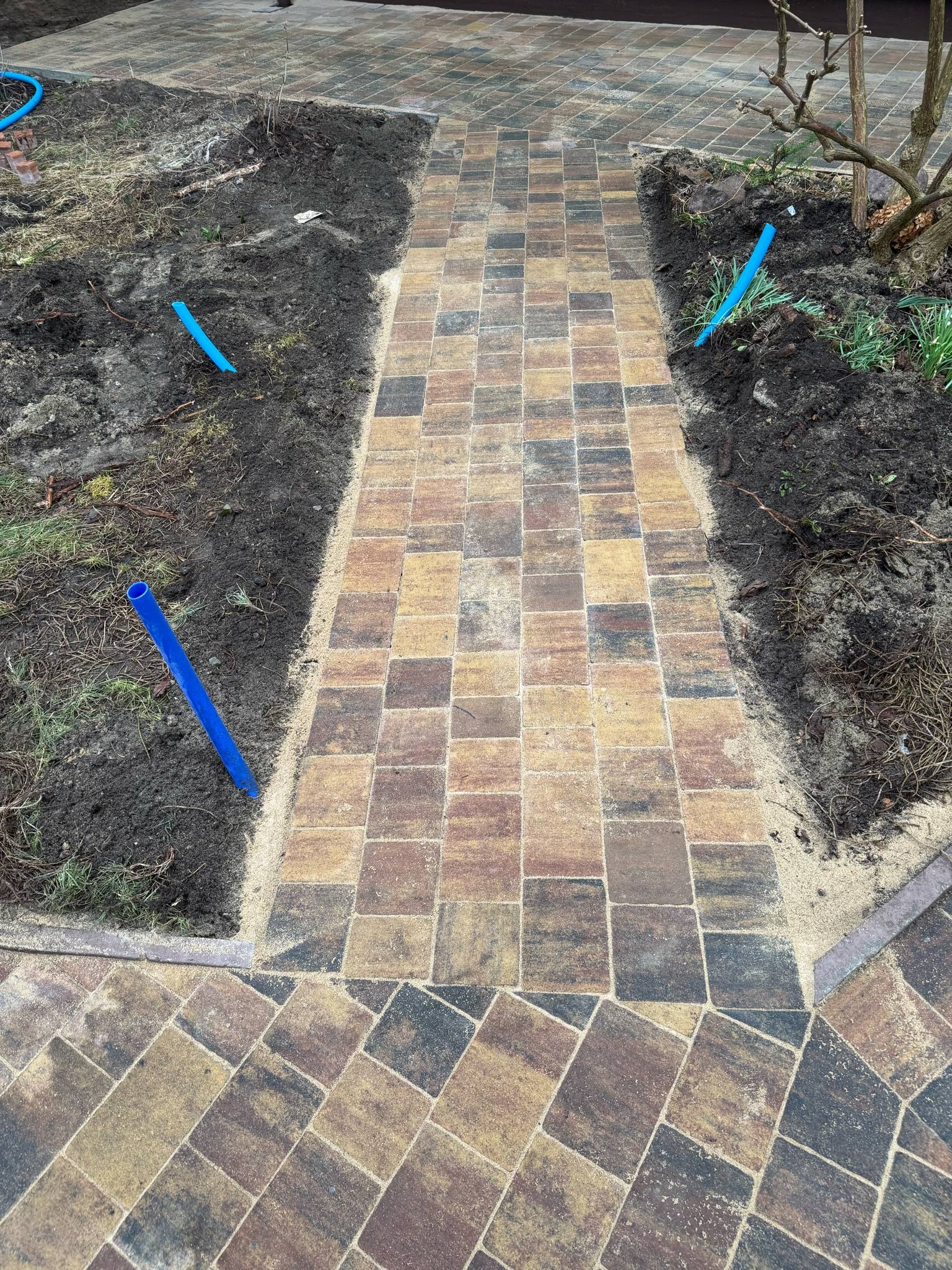 Ścieżka z kostki brukowej w odcieniach brązu i beżu, wyznaczona niebieskimi rurkami, prowadząca przez świeżo przygotowane rabaty ziemne. Widok z góry.