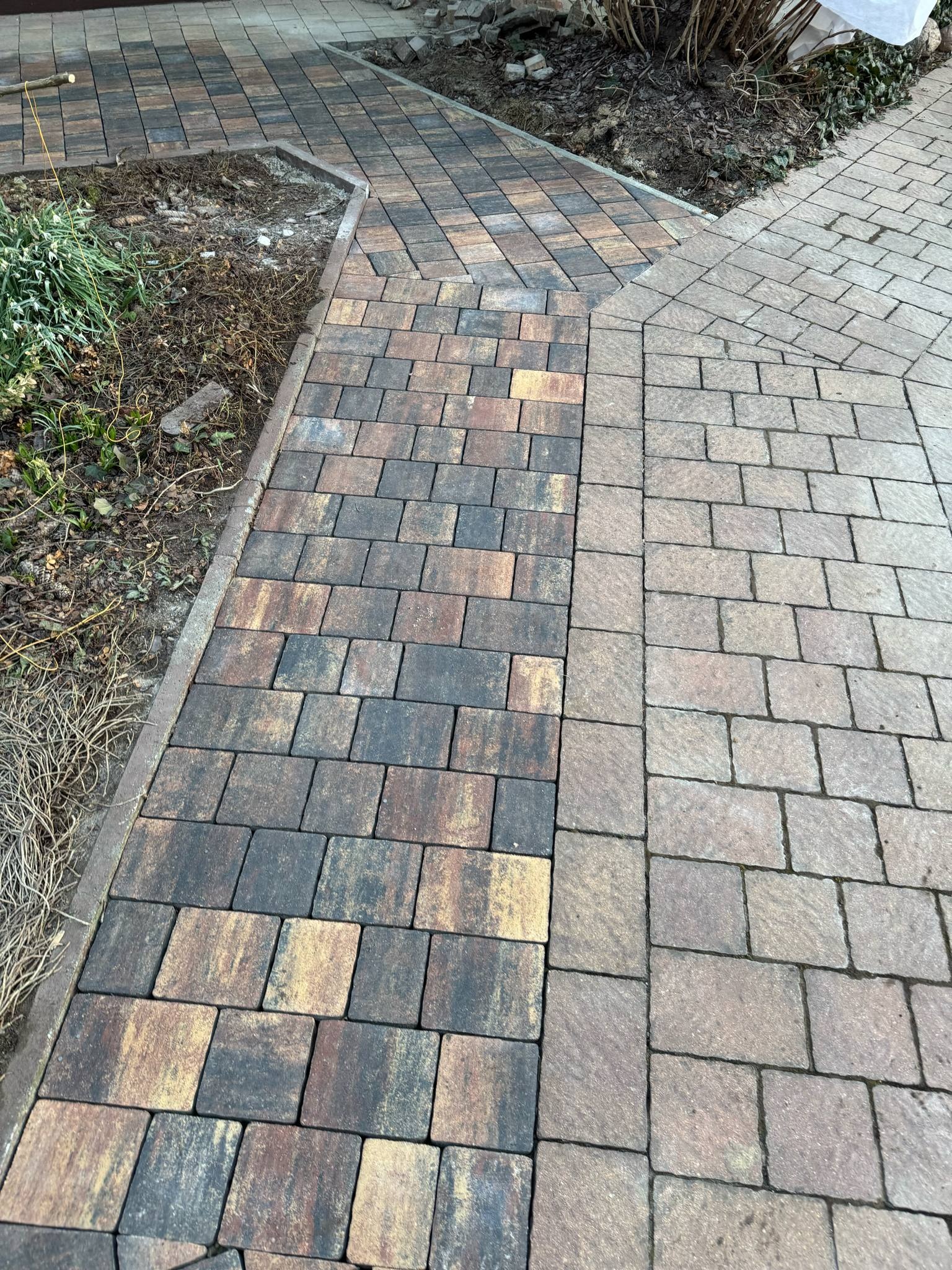 Ułożona kostka brukowa w dwóch odcieniach, tworząca ścieżkę i obrzeże rabaty. Widoczne różnice w kolorze kostki i wzorze ułożenia.