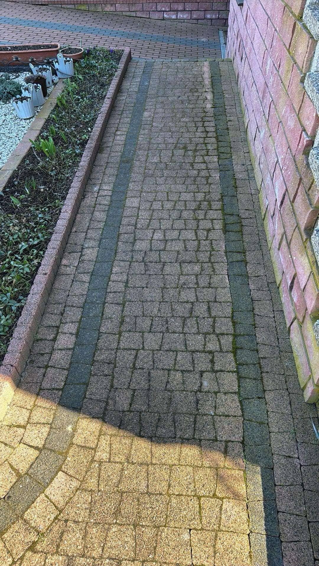 Ułożona kostka brukowa wzdłuż ściany, z fragmentem rabaty kwiatowej po lewej stronie. Widoczne ślady użytkowania i delikatny cień na części powierzchni.