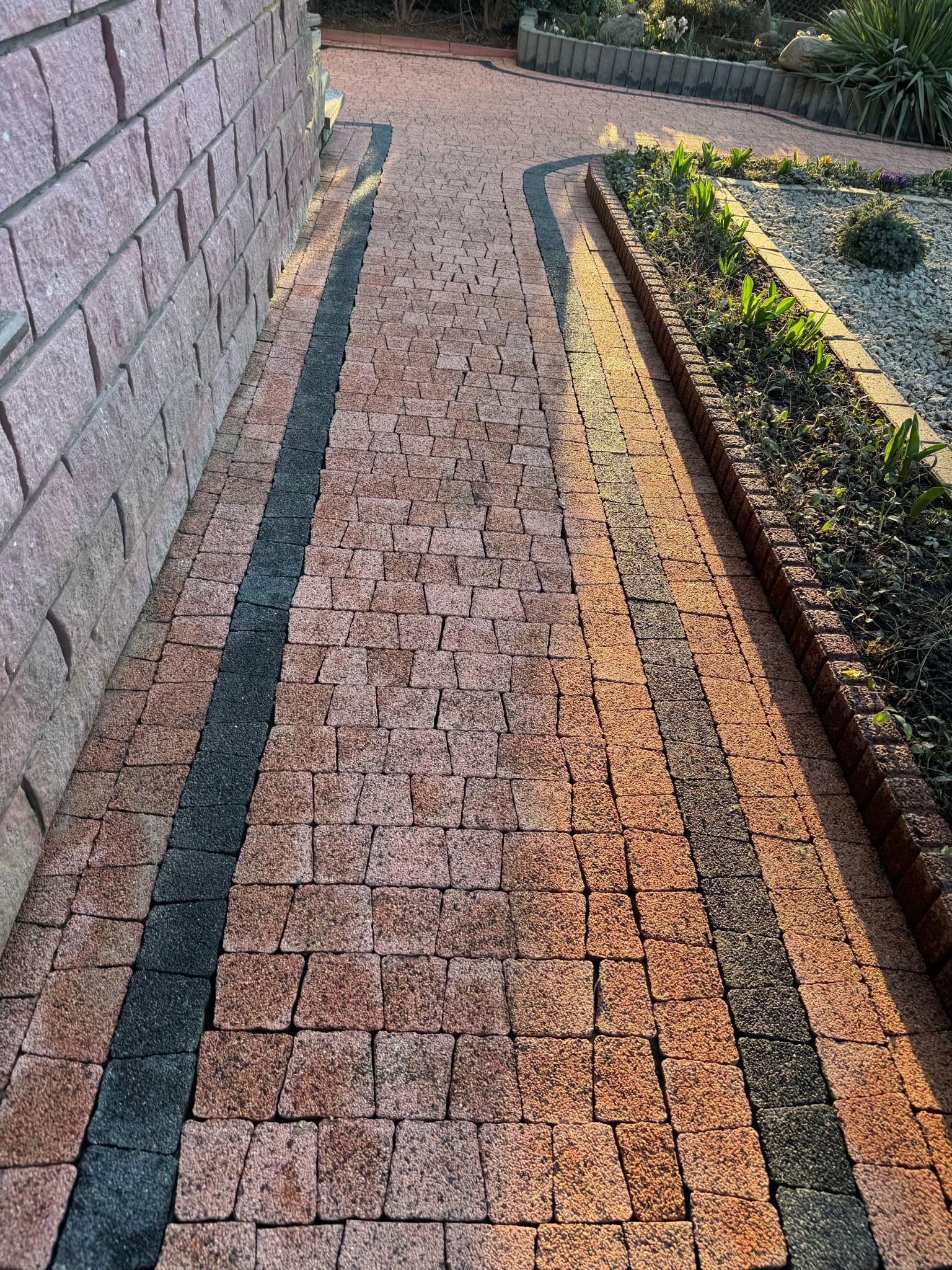 Kaskadowa ścieżka z kostki brukowej w odcieniach czerwieni i czerni, obrzeża rabat z roślinnością, mur z kamienia w tle, widok z góry, ciepłe światło.