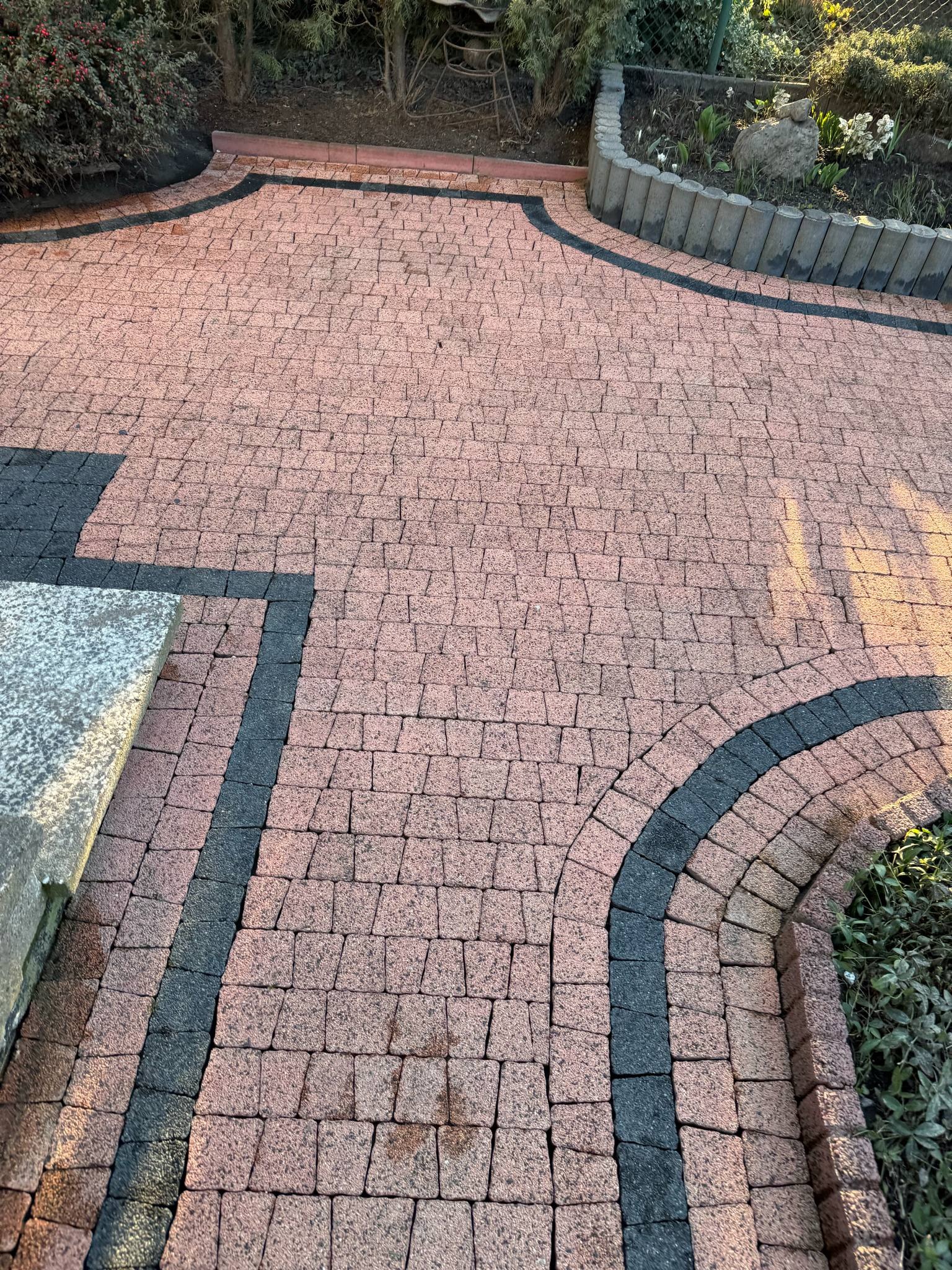 Kaskada różowo-czarnej kostki brukowej tworząca ścieżkę w ogrodzie, z widocznym obrzeżem i fragmentem rabaty kwiatowej. Ujęcie z góry, w ciepłym świetle słonecznym.