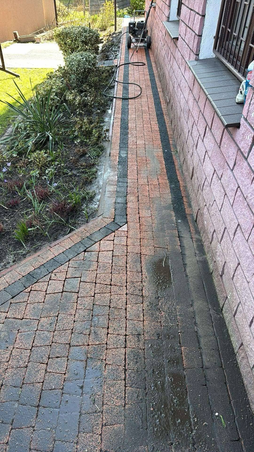 Częściowo wyczyszczona kostka brukowa w odcieniach czerwieni i czerni, obok muru budynku i rabaty z roślinami. Na końcu ścieżki myjka ciśnieniowa.