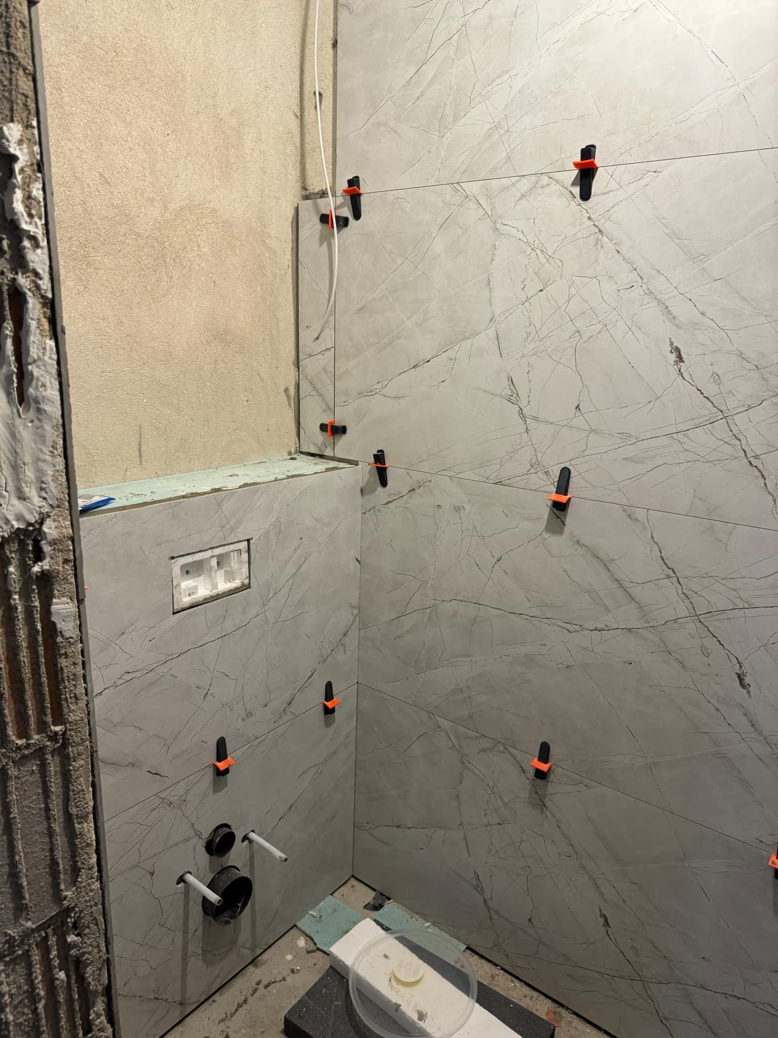 Ściana w trakcie wykańczania szarymi płytkami imitującymi marmur, z użyciem klipsów poziomujących. Widoczne przygotowane przyłącza wodne i fragment surowej ściany.