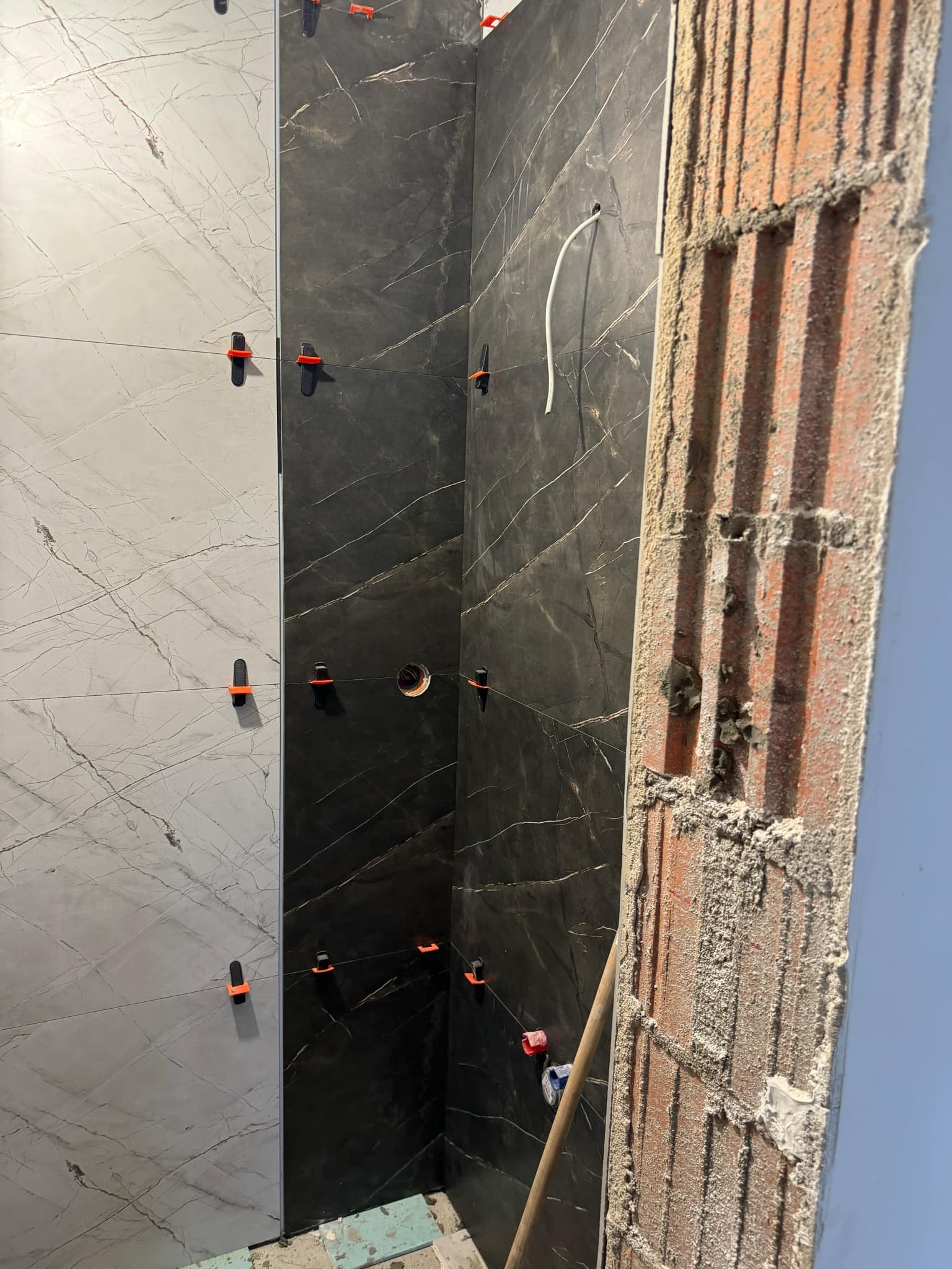 Wnętrze w trakcie wykańczania: częściowo położone marmurowe płytki w odcieniach szarości i czerni, widoczne pomarańczowe klipsy poziomujące, surowa cegła z zaprawą.