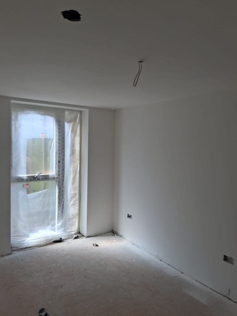 Wnętrze pokoju po tynkowaniu ścian na biało, okno zabezpieczone folią, widoczne gniazdka elektryczne i otwór w suficie na oświetlenie.
