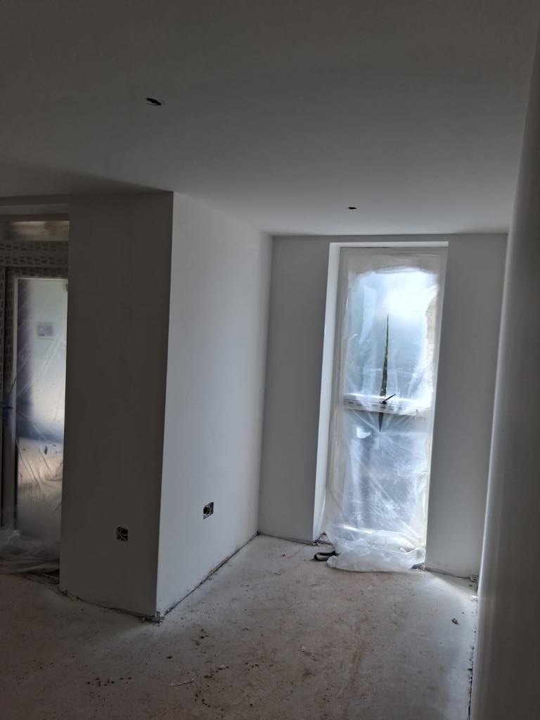 Białe, świeżo pomalowane wnętrze pokoju z oknem zabezpieczonym folią malarską; widoczne gniazdka elektryczne w ścianie i surowa podłoga.