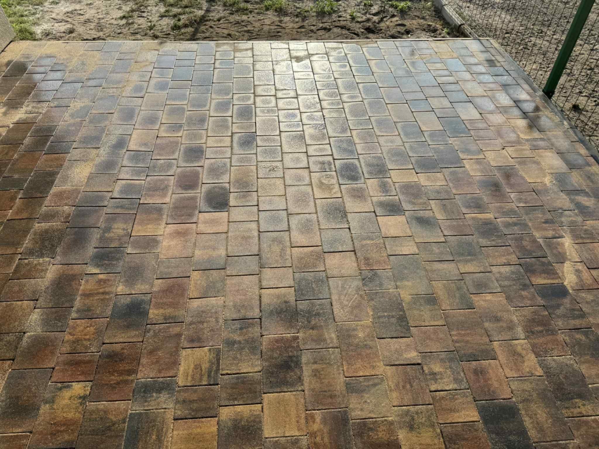 Ułożona kostka brukowa w odcieniach brązu i szarości, widok z góry. Kostka prostokątna, ułożona równo, z piaskiem w szczelinach. Fragment trawnika w tle.