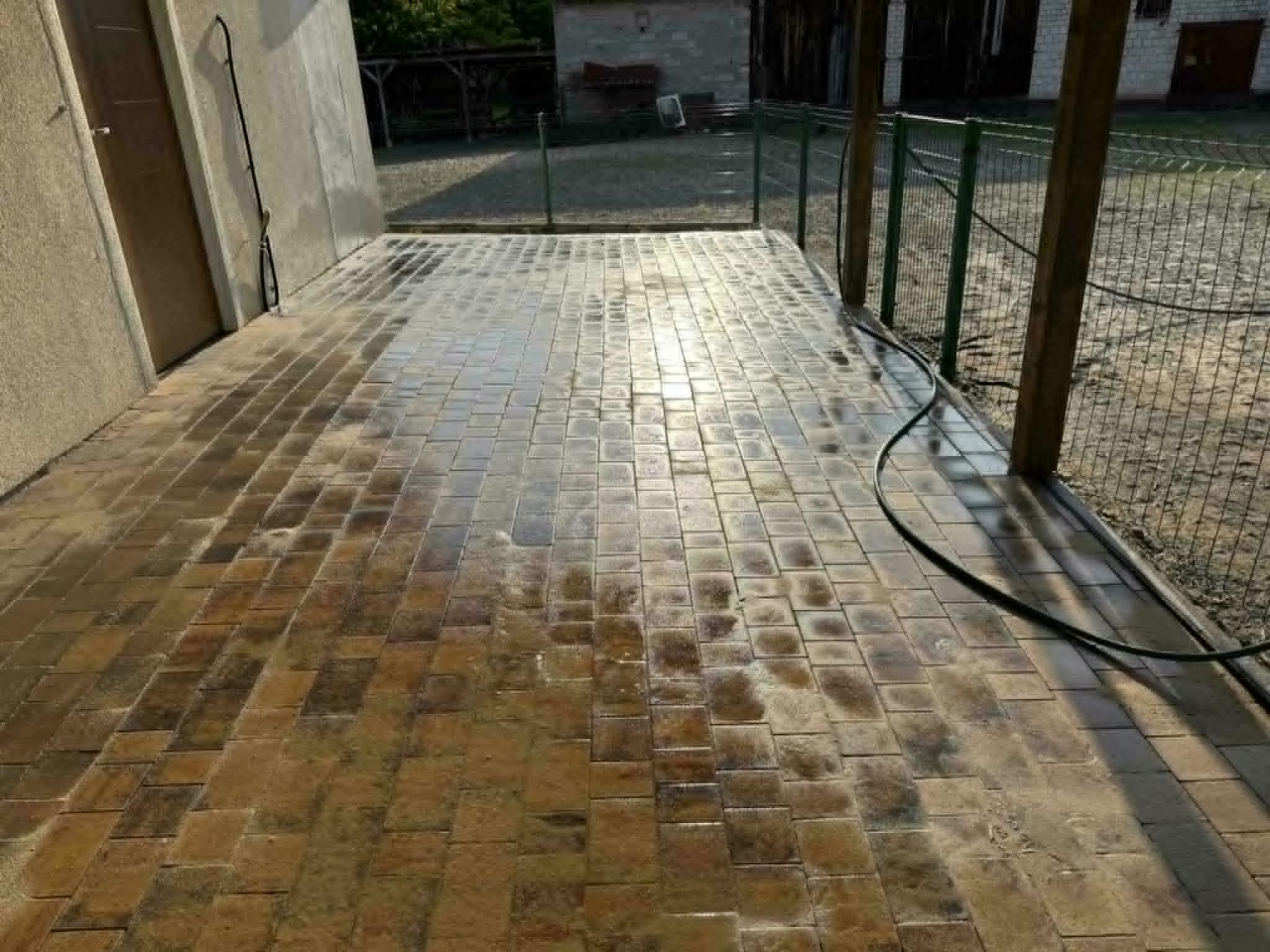Brązowa kostka brukowa po czyszczeniu, mokra powierzchnia odbija światło słoneczne. W tle zielona siatka ogrodzeniowa i budynek.