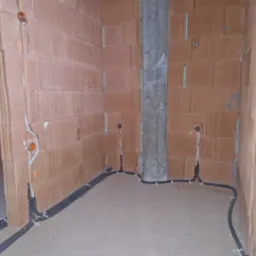 Surowy stan wnętrza domu w trakcie prac elektrycznych: widoczne kable w peszlach prowadzone po podłodze i pionowo po ścianach z pustaków ceramicznych, zakończone puszkami instalacyjnymi.