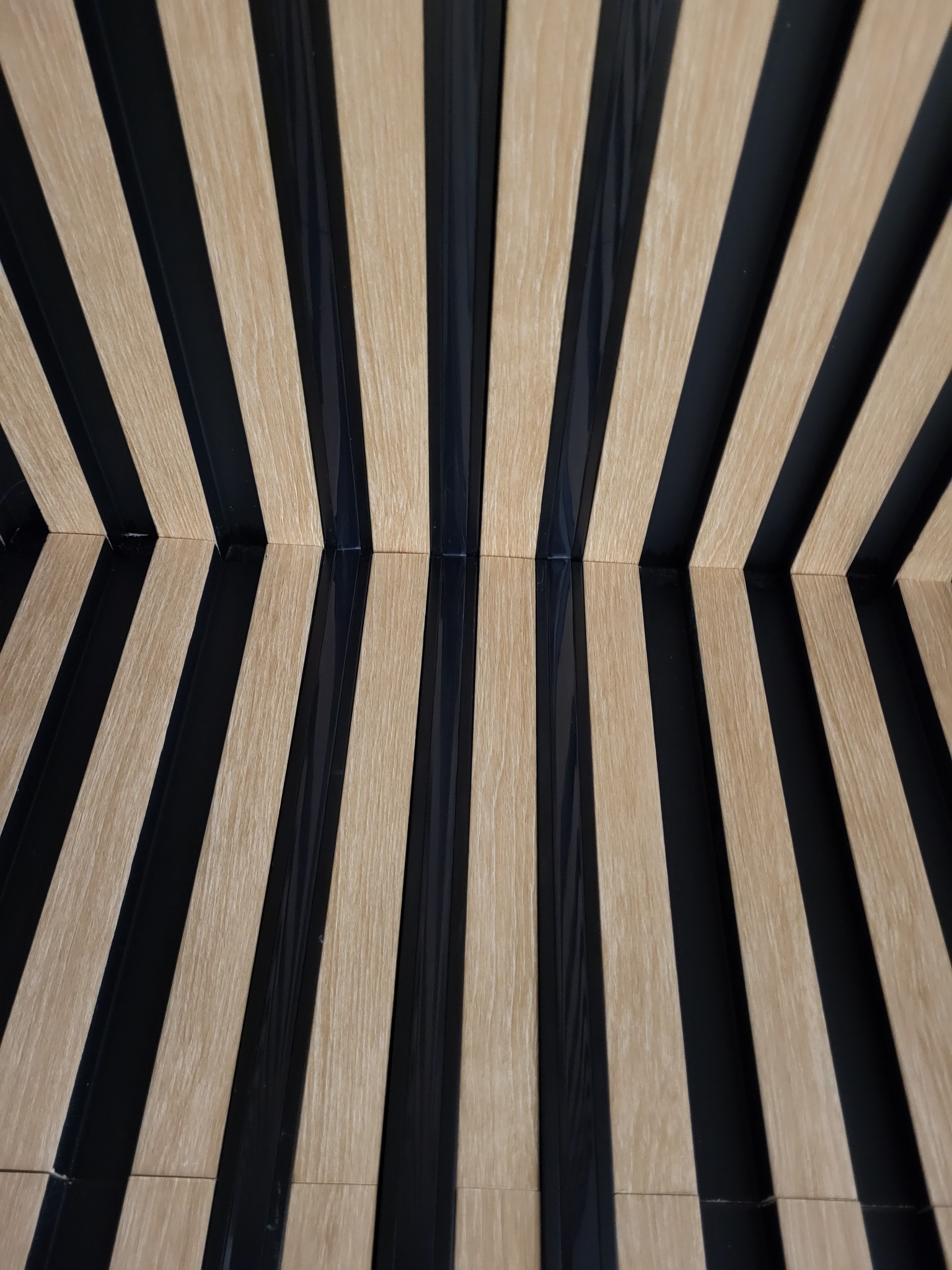 Ukryte oświetlenie LED, zamontowane pomiędzy drewnianymi lamelkami w tonacji jasnego drewna i czarnych listew, tworzące dekoracyjny wzór na ścianie.