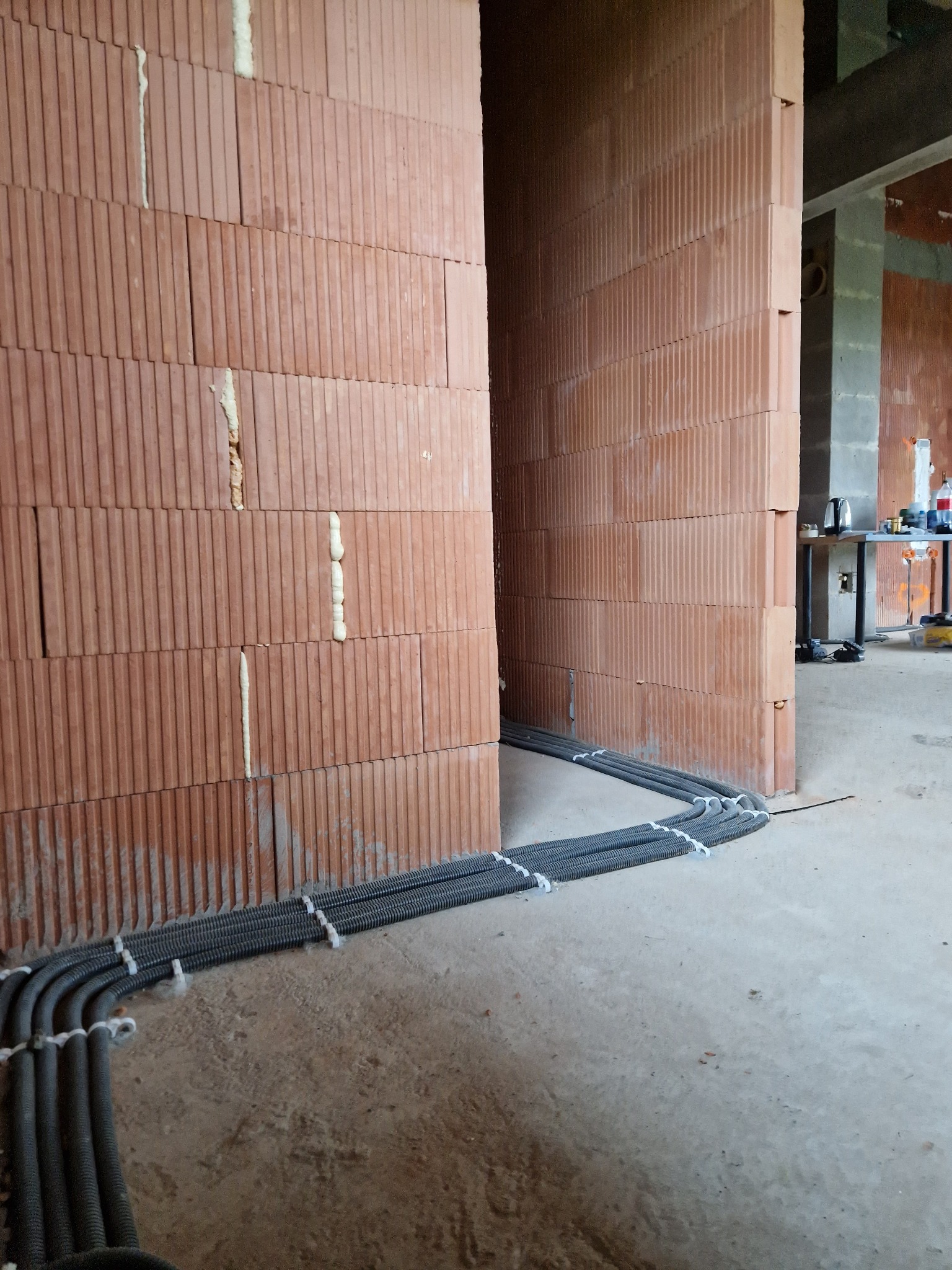 Kable elektryczne w peszlach, ułożone wzdłuż ściany z czerwonej cegły dziurawki na surowym betonie, zabezpieczone białymi opaskami zaciskowymi w trakcie budowy domu.