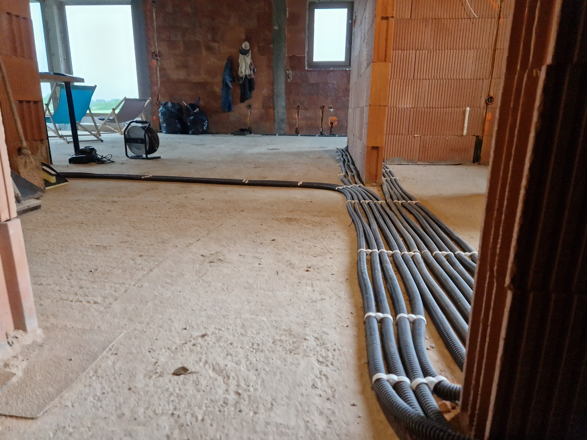 Wiązka przewodów elektrycznych w karbowanych rurach, ułożona na surowej posadzce w trakcie prac instalacyjnych w budynku z czerwonej cegły. W tle widoczne okno, krzesła plażowe i lampa halogenowa.
