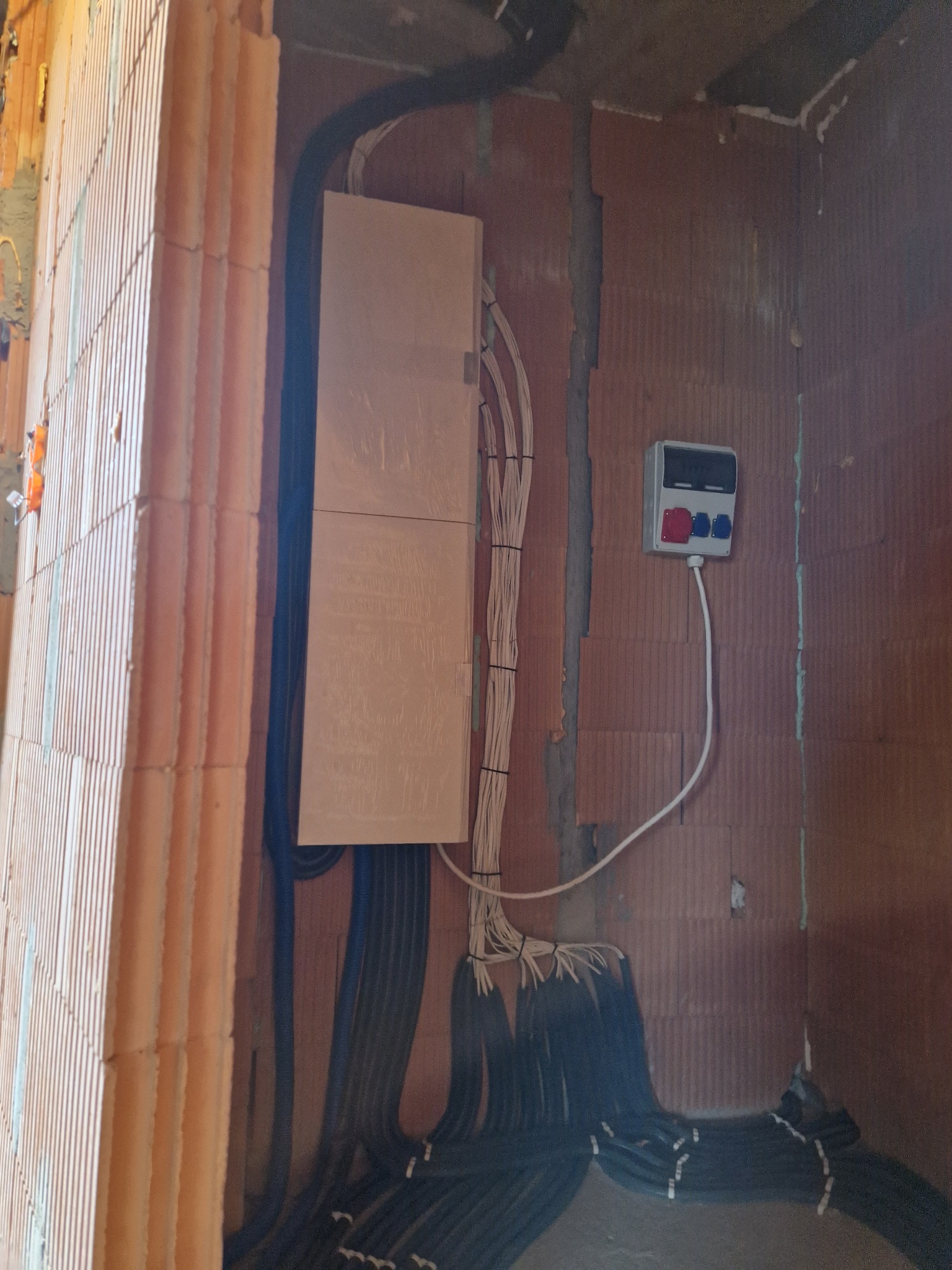 Rozdzielnia elektryczna z licznymi przewodami w peszlach, zamontowana w surowym wnętrzu domu z cegły.