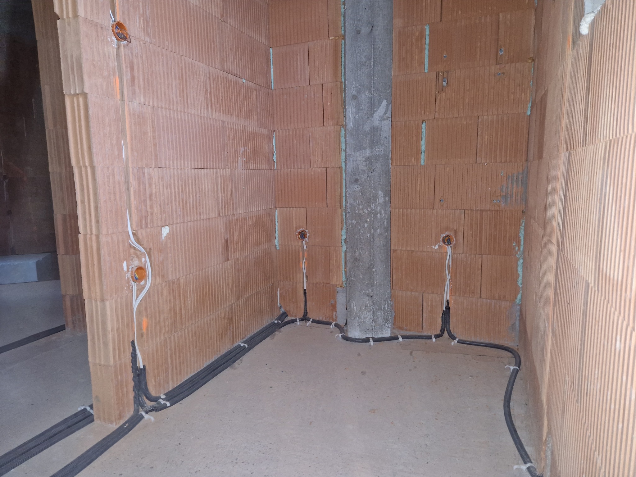 Surowy stan wnętrza domu w trakcie prac elektrycznych: widoczne kable w peszlach prowadzone po podłodze i pionowo po ścianach z pustaków ceramicznych, zakończone puszkami instalacyjnymi.