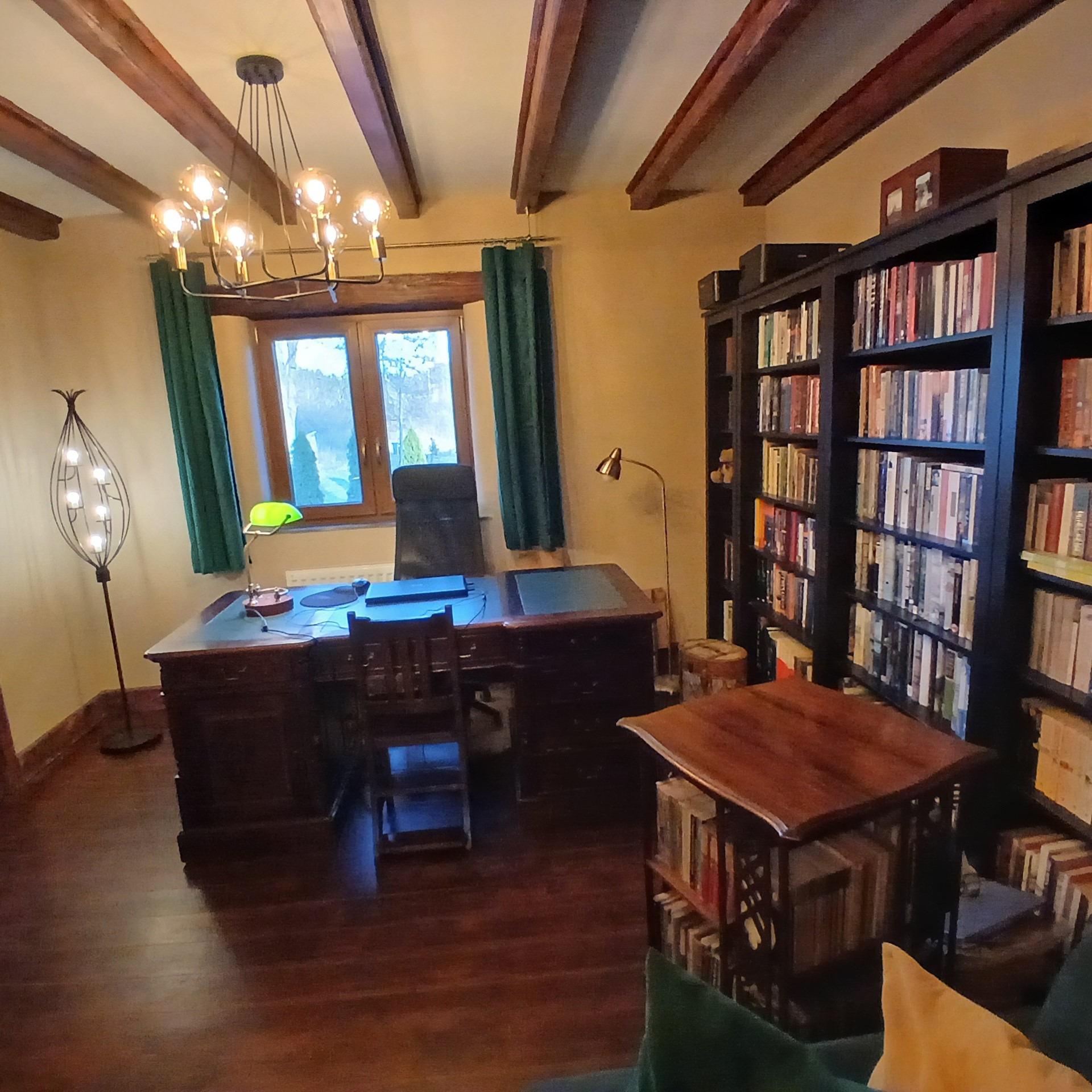 Gabinet z ciemnym drewnianym biurkiem, biblioteczką i zielonymi zasłonami. Widoczna lampa i okno z widokiem na zewnątrz. Ciepłe oświetlenie wnętrza.