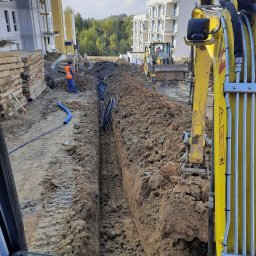 JAKOP Budownictwo ziemne i kubaturowe JAKUB DUCZMIŃSKI - Głęboki wykop w ziemi z ułożonymi rurami i kablami, widoczny pracownik w pomarańczowej kamizelce, koparka oraz budynki mieszkalne w tle, wykonane z perspektywy kabiny maszyny.
