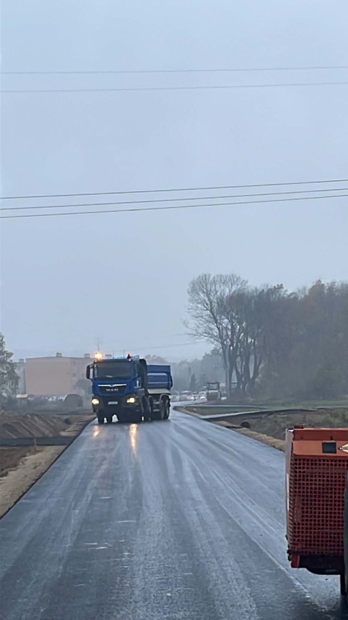 Nawierzchnia świeżo wylana, mokra droga z widocznym ciężkim sprzętem budowlanym, w tym ciężarówka MAN i walec, w tle drzewa i budynek w pochmurny dzień.