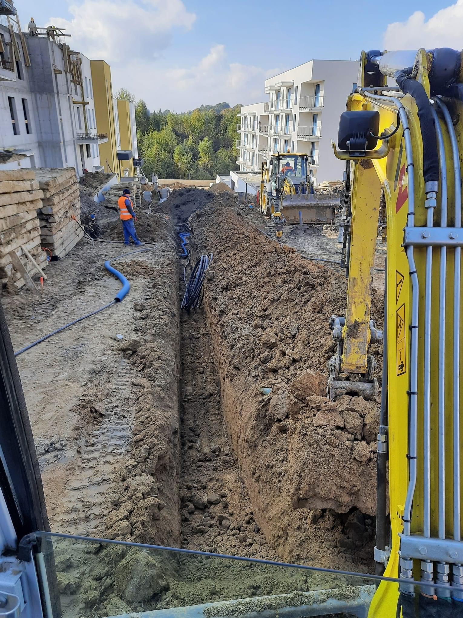 Głęboki wykop w ziemi z ułożonymi rurami i kablami, widoczny pracownik w pomarańczowej kamizelce, koparka oraz budynki mieszkalne w tle, wykonane z perspektywy kabiny maszyny.