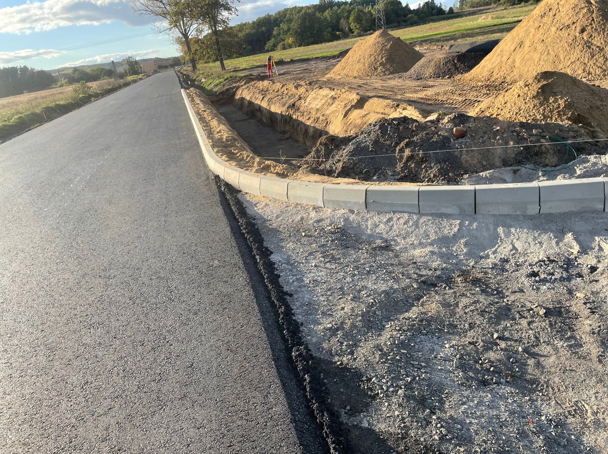 Budowa drogi: świeżo położony asfalt sąsiaduje z wykopem zabezpieczonym betonowymi krawężnikami, w tle pryzmy piasku i geodeta z niwelatorem.