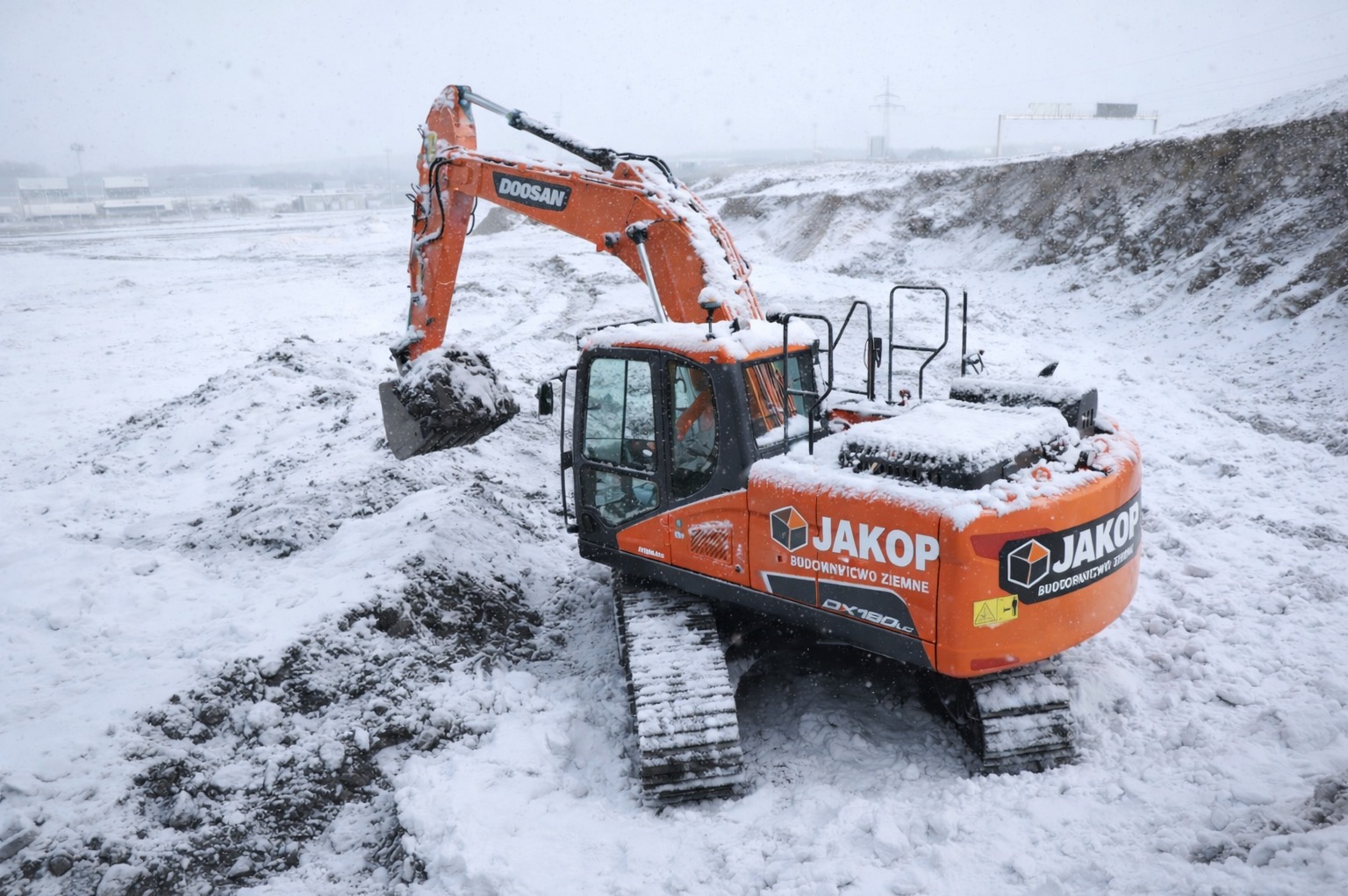 Pomarańczowa koparka Doosan JAKOP w zimowej scenerii, przysypana śniegiem, z łyżką pełną ziemi. W tle zasypane śniegiem pole i linia energetyczna.