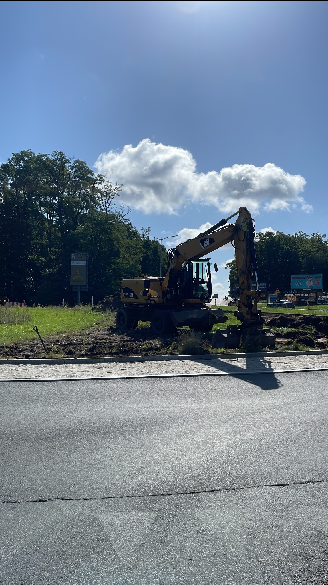 Żółta koparka CAT na tle zieleni i błękitnego nieba z chmurami, wykonująca prace ziemne przy krawężniku. Widoczny fragment drogi asfaltowej.