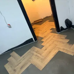 Układanie paneli podłogowych w jodełkę na szarym podłożu, widoczne niedociągnięcia przy ścianach, przygotowanie do listew przypodłogowych.