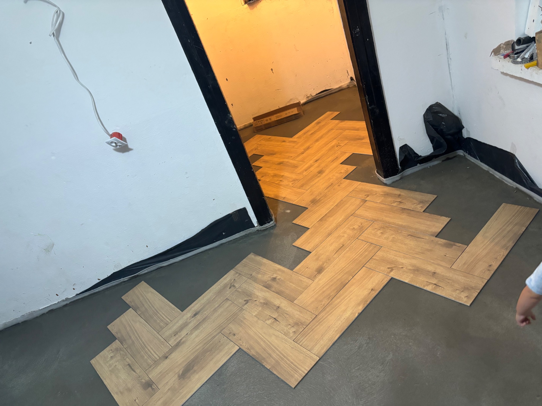 Układanie paneli podłogowych w jodełkę na szarym podłożu, widoczne niedociągnięcia przy ścianach, przygotowanie do listew przypodłogowych.