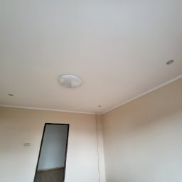 Pan Robótka - Pomalowane na beżowo ściany i biały sufit z zamontowanym okrągłym plafonem i dwoma małymi punktowymi lampkami halogenowymi, widoczne listwy przypodłogowe i ościeżnica drzwi.