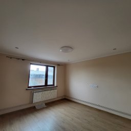 Pan Robótka - Pusty pokój po remoncie z panelami podłogowymi, grzejnikiem pod oknem i gładkimi, pomalowanymi ścianami w odcieniach beżu. Widoczna listwa przypodłogowa i instalacja elektryczna.