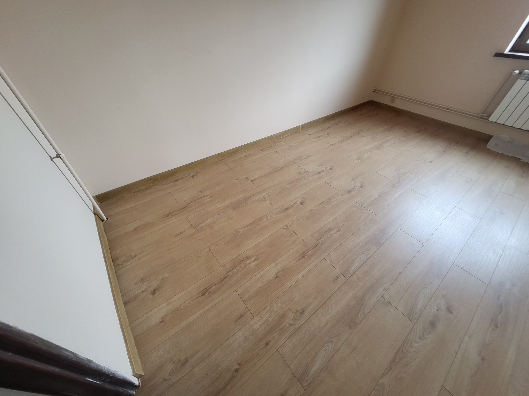 Jasne panele podłogowe ułożone w pustym pokoju z grzejnikiem pod oknem i świeżo pomalowanymi ścianami.