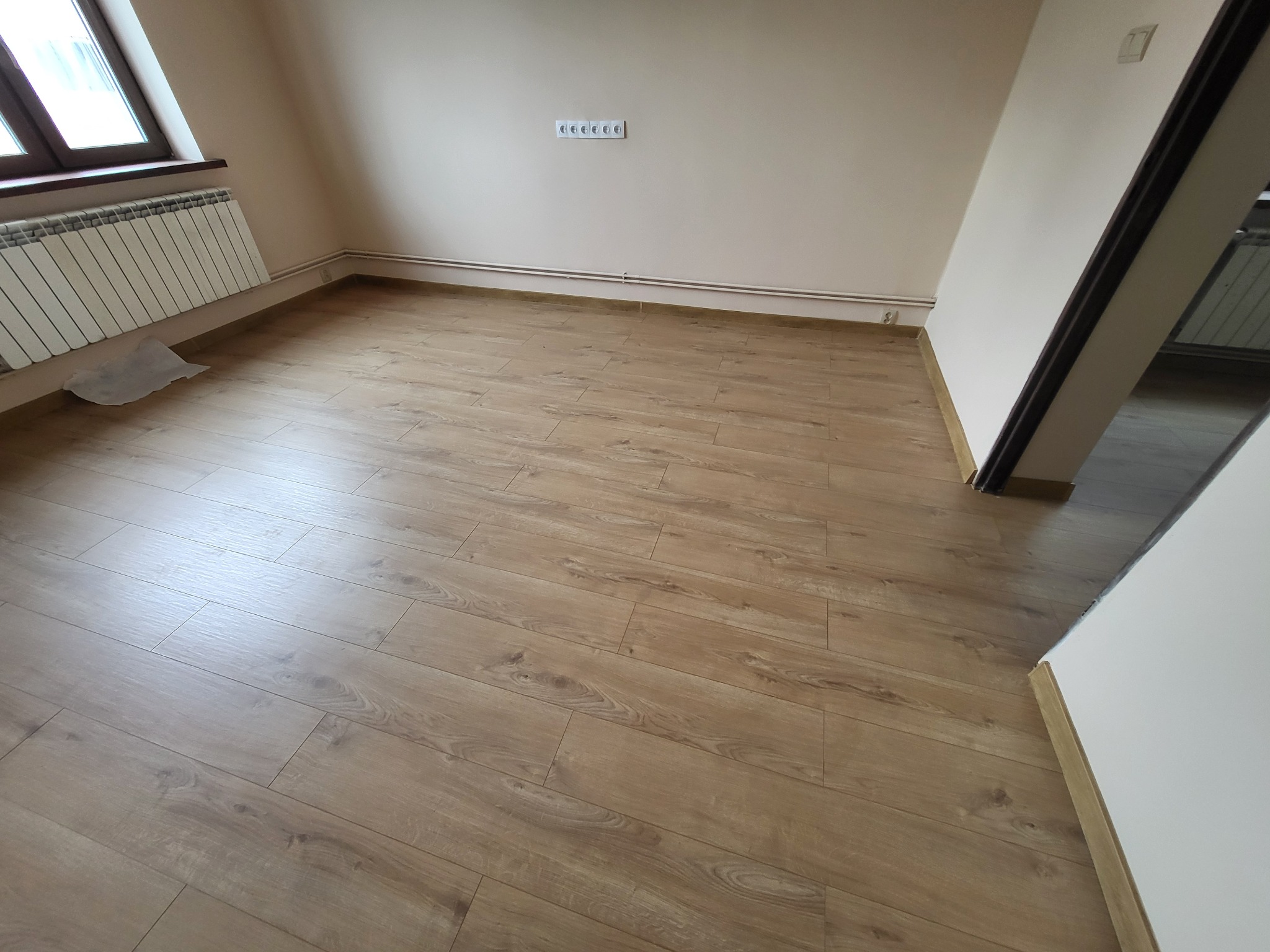 Jasne panele podłogowe imitujące drewno, ułożone w pustym pokoju z grzejnikiem pod oknem i białymi ścianami.