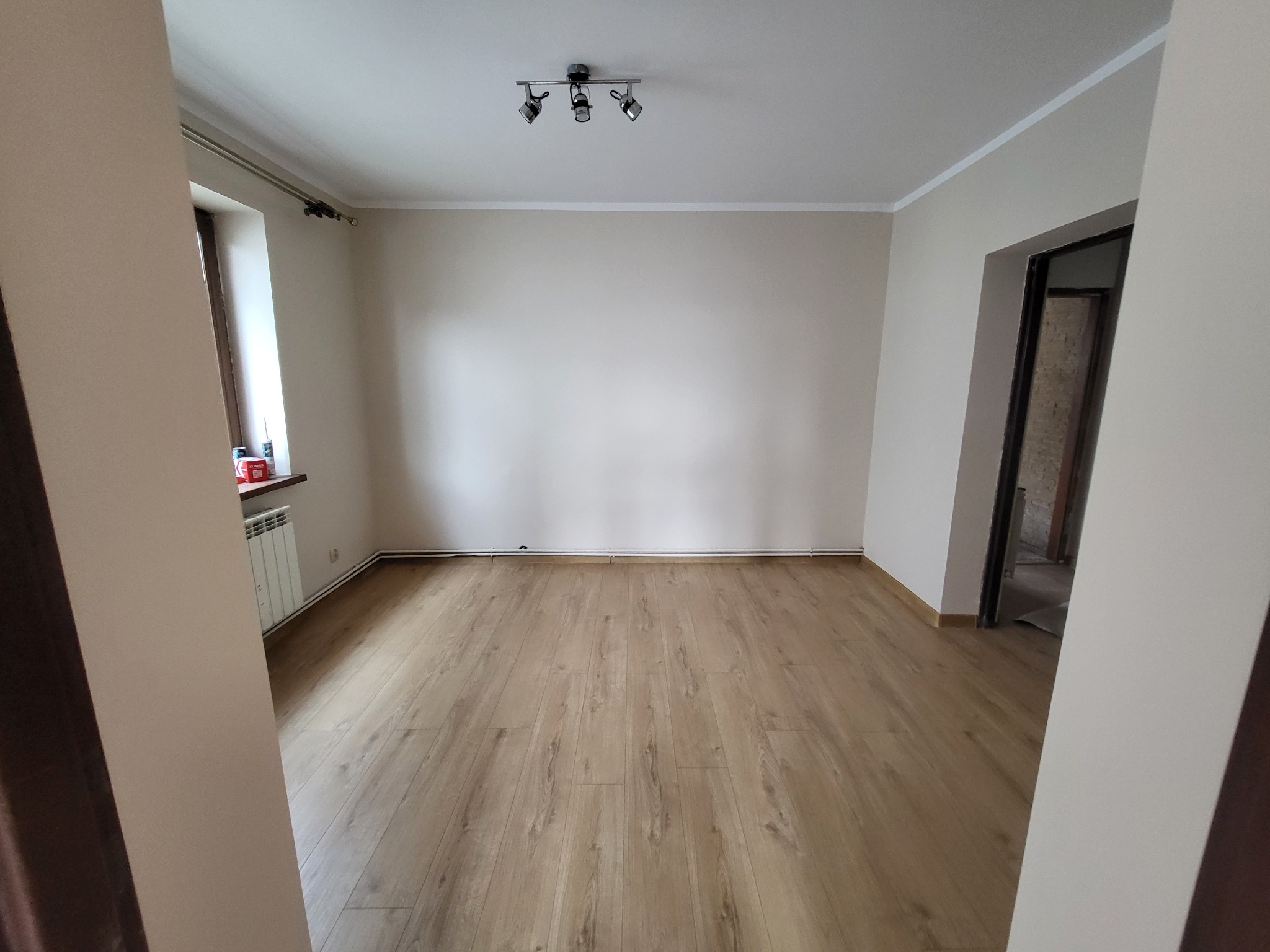 Jasne panele podłogowe ułożone w pustym pokoju w Czeladzi, świeżo po remoncie, widok z perspektywy wejścia, ściany pomalowane na jasny beż, widoczny grzejnik i listwy przypodłogowe.